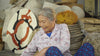 Elderly artisan smiling while surrounded by beautifully handcrafted woven baskets and traditional decor pieces.