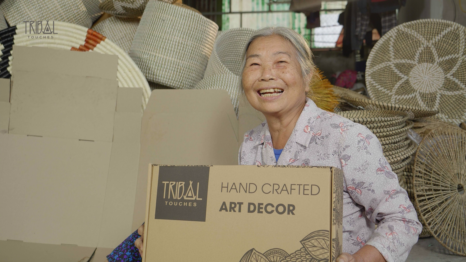 Grandma Lan holding a Tribal Touches handcrafted art decor box, surrounded by artisan baskets.